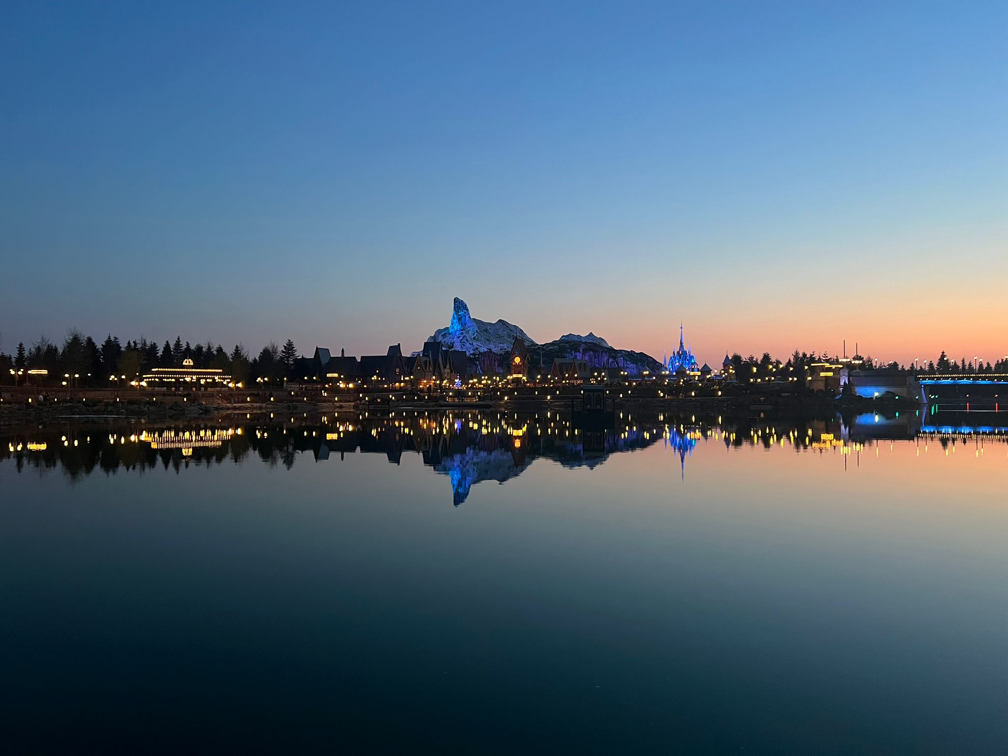 Dusk over Lake Disney, with the lit mountain of World of Frozen and the park's castle reflected in still water